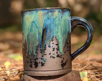 May include: A ceramic mug with a blue, green, and black glaze. The mug has a silhouette of a forest scene on the front.