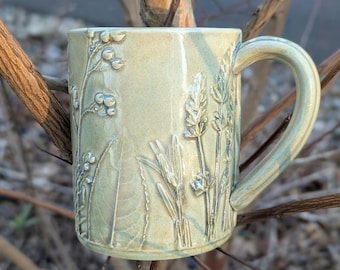 May include: A handmade ceramic mug with a light green glaze. The mug features embossed botanical designs of various plants and flowers. The handle is curved and integrated into the mug's design. The mug is a unique piece of functional art.