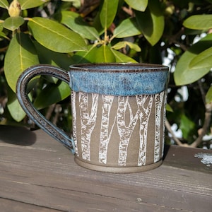 May include: A ceramic mug with a brown and blue glaze. The mug has a white design of birch trees on the front.