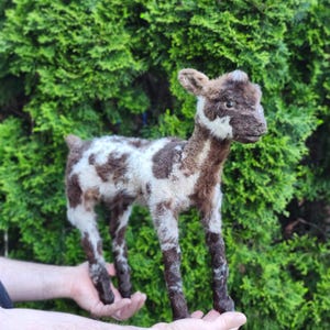 May include: A handcrafted lamb figurine with a brown and white spotted pattern. The lamb is standing, with its legs and head in a darker brown color. The figurine is held in front of a green bush.