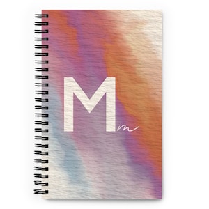 May include: A spiral bound notebook with a watercolor-style cover featuring a large white letter "M" and a smaller handwritten "m" in the bottom right corner. The background is a blend of orange, pink, purple, and blue hues.