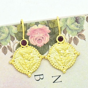 May include: Gold-coloured earrings with a square, ornate design. Each earring has a small, round, red gemstone at the top and a detailed relief image within the square. The earrings are displayed on a floral-patterned surface.