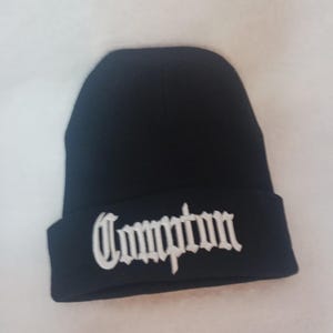 May include: Black beanie with the word "Compton" embroidered in white, featuring a classic, folded cuff. The hat is made of a soft, knit material.