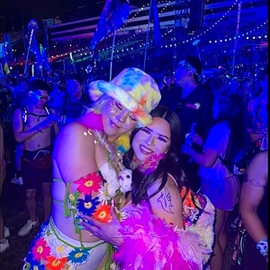 May include: Two people are posing for a photo at a music festival. The person on the left is wearing a white crocheted hat with a rainbow tie-dye pattern, a white crocheted top with colorful flowers, and a white skirt with colorful flowers. The person on the right is wearing a pink feather boa and a black tank top with a white bird tattoo on their arm.