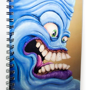 May include: Spiral-bound notebook featuring a vibrant, cartoonish monster face. The creature has a blue complexion, large white teeth, and a purple mouth. The notebook's design is detailed and eye-catching, perfect for creative writing or sketching.