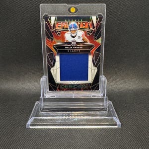 May include: A sports trading card featuring Malik Nabers of the Giants, labeled "Rookie" and "Obsidian." The card has a blue jersey swatch and is encased in a clear protective holder, displayed on a clear stand.