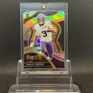 May include: A Jordan Addison football trading card in a protective case. The card features a player in a white and purple uniform with the number 3. The card is labeled "Select" and "Vikings". The background has a holographic design.