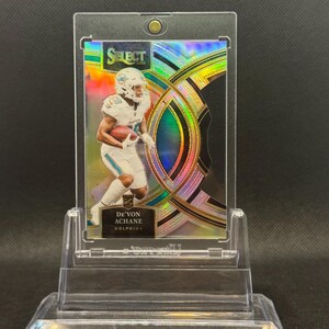 May include: A football trading card featuring De'Von Achane of the Dolphins. The card is encased in a clear plastic holder and displayed on a clear stand. The card has a holographic design with the word "SELECT" at the top.