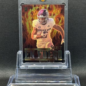 May include: A clear plastic card holder with a football trading card inside. The card features a football player in a maroon and white uniform with the text "Texas A&M" on the front. The card has a holographic background with flames and the text "En Fuego" in gold lettering.