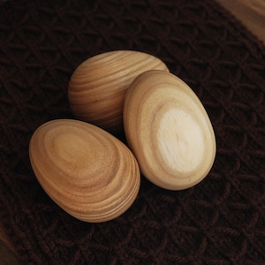 May include: Three wooden eggs with a natural wood grain finish. The eggs are arranged on a brown knitted surface.