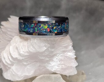 May include: A silver-colored ring with a band of colorful, iridescent material. The ring's band features a mosaic of blue, green, red, and yellow fragments. The ring is displayed on a white, textured surface.
