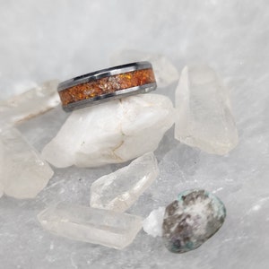 May include: A close-up of a ring with a dark gray band and a center of orange and white speckled material. The ring is resting on white quartz crystals and a small, speckled stone.