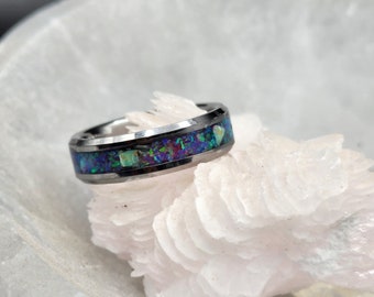 May include: A silver-toned ring with a band of inlaid, colorful opal. The opal features a mix of blue, green, and purple hues. The ring is set against a white, textured background, highlighting the jewelry's design.
