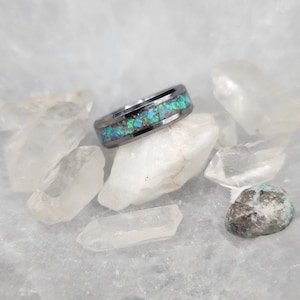 May include: A silver-colored ring with a band of inlaid, iridescent blue and green stones. The ring is set against a backdrop of clear quartz crystals and a white, marble-like surface.