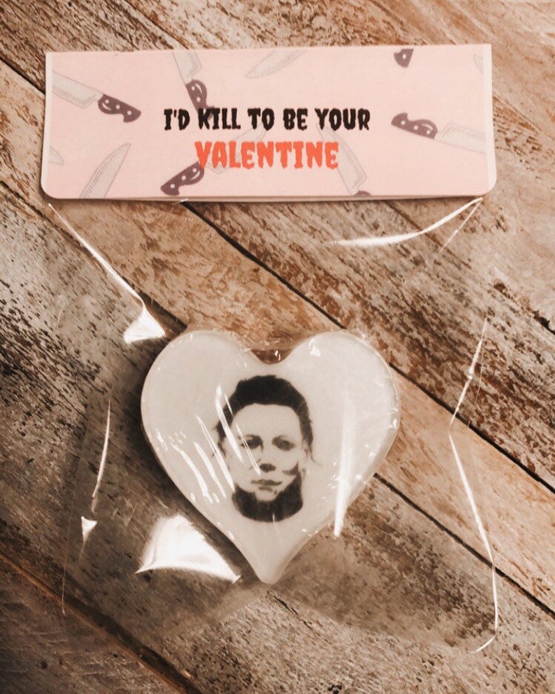 Op de afbeelding: Een witte hartvormige koek met een zwart-witte afbeelding van Michael Myers uit de Halloween-films. De koek is verpakt in een doorzichtige plastic zak met een roze kaart waarop staat "Ik zou alles doen om je Valentijn te zijn" met een mespatroon.