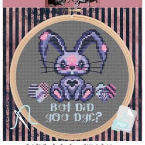 Può includere: Uno schema di punto croce con un coniglio viola con un pentagramma sulla fronte, che tiene un cuore e un gomitolo di lana. Il testo "But Did You Dye?" è sotto il coniglio. Lo schema è incorniciato in un cerchio di legno e ha uno sfondo a strisce rosa e nero.