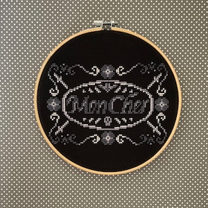 May include: Black and white embroidery hoop with a black background and white stitching. The stitching depicts a vintage-style logo with the text "Mon Cheri" in a script font.