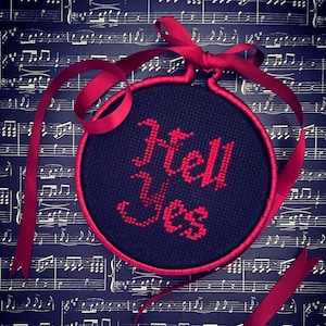 May include: A round black embroidery hoop with a red embroidered phrase "Hell Yes" on a black background. The hoop is tied with a red ribbon.