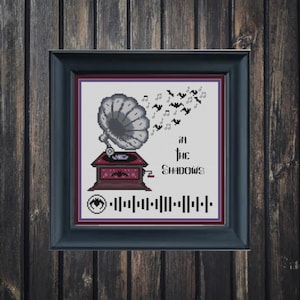 以下が含まれることがあります： 黒いフレームに入ったクロスステッチアート。デザインは、音符とコウモリが付いたヴィンテージ蓄音機が特徴です。「in the Shadows」というテキストとバーコード、コウモリのシンボルがあります。