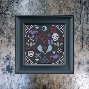 May include: A framed cross-stitch featuring a dark background with various Halloween-themed motifs. The design includes skulls, bats, ghosts, crosses, and the number 13. The frame is a dark gray color.