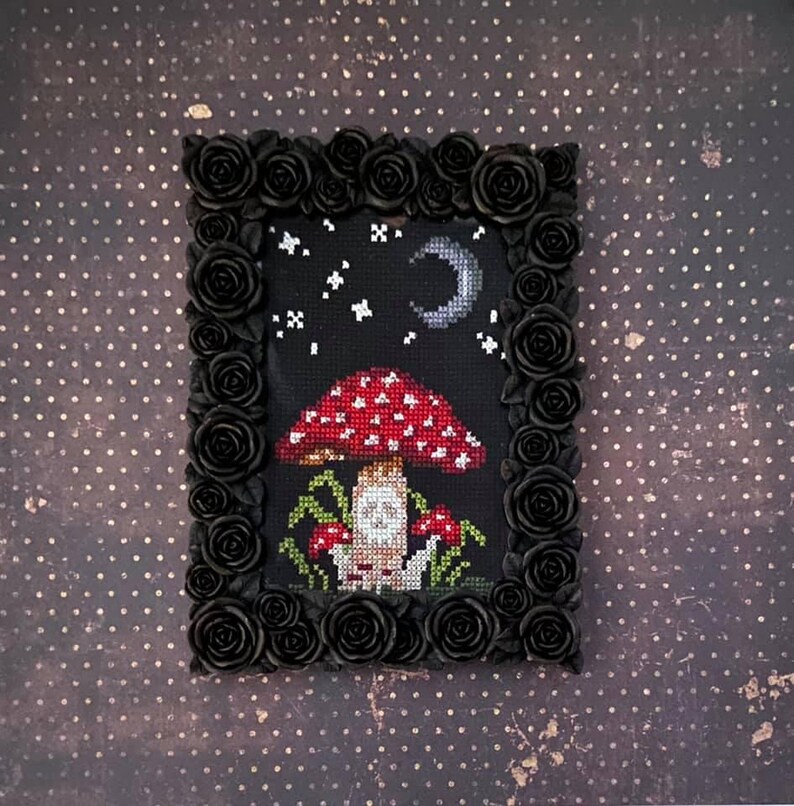 May include: A cross-stitch embroidery framed in black with a black rose border. The embroidery depicts a red and white mushroom with a white face under a crescent moon and stars.