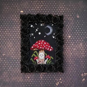 May include: A cross-stitch embroidery framed in black with a black rose border. The embroidery depicts a red and white mushroom with a white face under a crescent moon and stars.