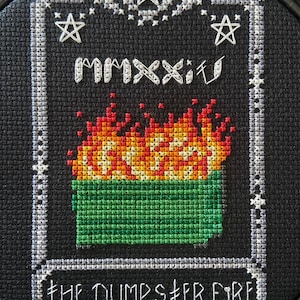 Peut inclure: Broderie au point de croix sur tissu noir, représentant une poubelle verte en feu avec des flammes rouges, oranges et jaunes. Le texte "THE DUMPSTER FIRE" est brodé en dessous. L'année "MMXXII" est brodée au-dessus.