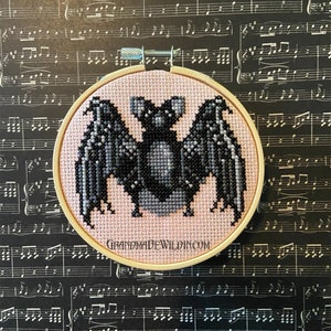 May include: A pink and black embroidered bat on a white background in a wooden embroidery hoop. The hoop is on a black and white sheet music background. The text "GRANDMADEWILDIN.COM" is on the hoop.