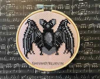 May include: A pink and black embroidered bat on a white background in a wooden embroidery hoop. The hoop is on a black and white sheet music background. The text "GRANDMADEWILDIN.COM" is on the hoop.