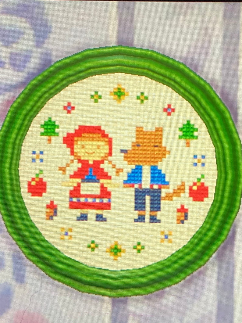 May include: A green framed embroidery hoop with a white background featuring a red-hooded girl and a wolf in blue clothing. The embroidery includes red apples, green trees, and yellow flowers.