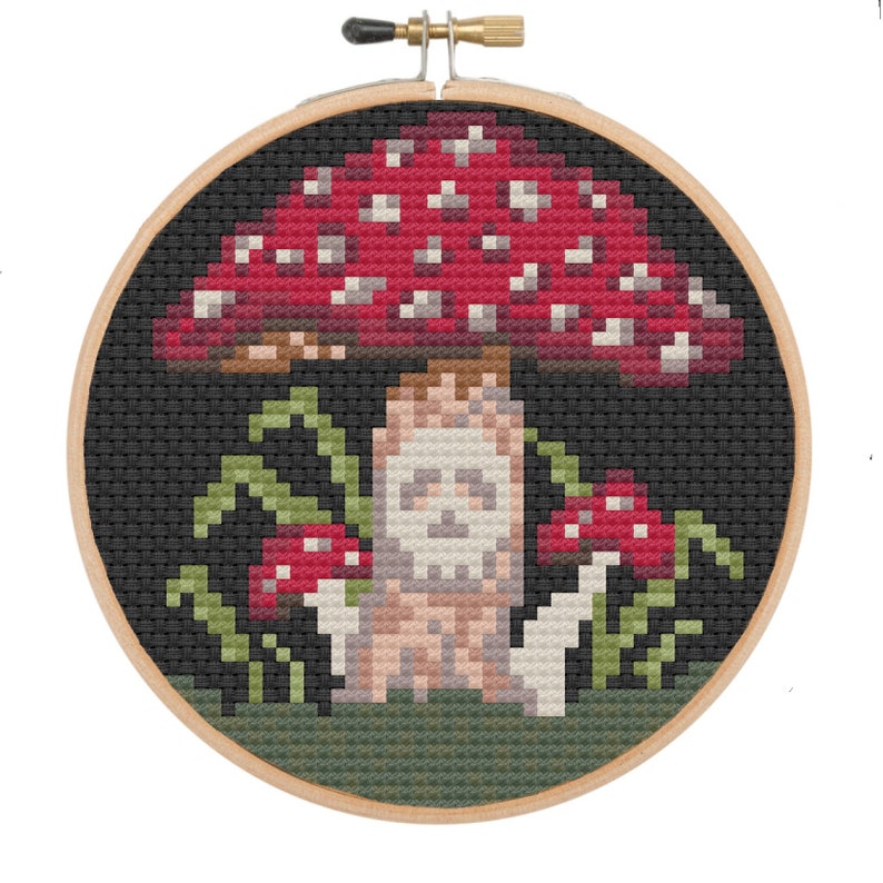 May include: Cross-stitch embroidery hoop featuring a red and white mushroom with a skull face on the stem. The mushroom is surrounded by green leaves and two smaller red mushrooms.