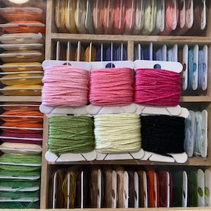 May include: A close-up of a wooden box filled with spools of embroidery floss. The spools are organized by color and are in a variety of shades, including pink, green, yellow, black, and brown.