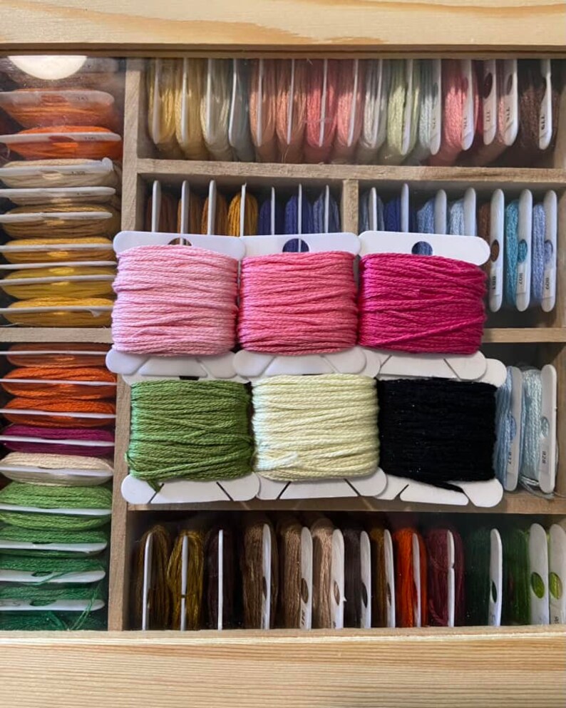 May include: A close-up of a wooden box filled with spools of embroidery floss. The spools are organized by color and are in a variety of shades, including pink, green, yellow, black, and brown.