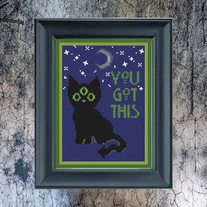 May include: Cross stitch pattern featuring a black cat with three green eyes, a crescent moon, and stars on a dark blue background. The text "YOU GOT THIS" is stitched in white.
