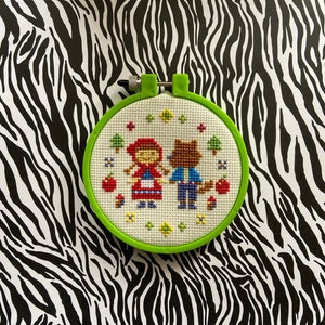 May include: A green embroidery hoop with a white cross-stitch design of Little Red Riding Hood and a wolf. The design features red, blue, yellow, and green thread.