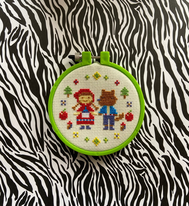 May include: A green embroidery hoop with a white cross-stitch design of Little Red Riding Hood and a wolf. The design features red, blue, yellow, and green thread.