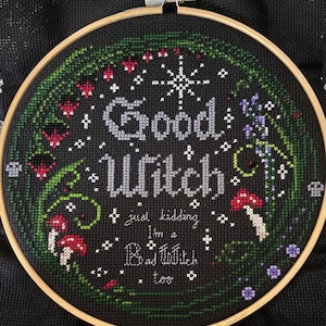 May include: Embroidered hoop art with a black background and white text that reads "Good Witch, just kidding I'm a Bad Witch too". The text is surrounded by green leaves, red flowers, and purple flowers.