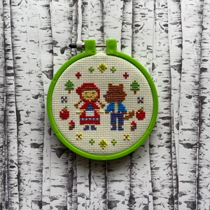 May include: A green embroidery hoop with a white fabric inside. The fabric has a cross-stitch design of Little Red Riding Hood and the wolf.