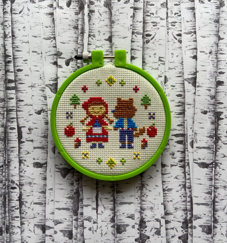 May include: A green embroidery hoop with a white fabric inside. The fabric has a cross-stitch design of Little Red Riding Hood and the wolf.