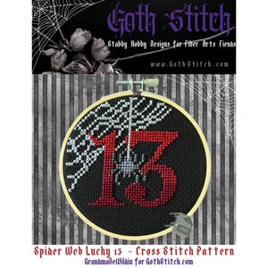May include: A black and white cross stitch pattern of a spider web with a red number 13 in the centre. The spider web is white and the spider is grey. The number 13 is red and the background is black. The pattern is in a round hoop. The text "Spider Web Lucky 13 - Cross Stitch Pattern" is below the hoop.
