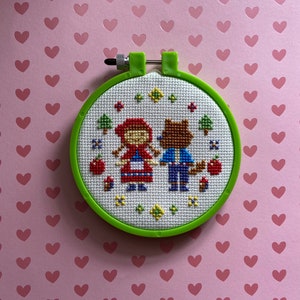 May include: A green embroidery hoop with a white cross-stitch design of Little Red Riding Hood and a wolf. The design is surrounded by red apples, green trees, and yellow flowers.