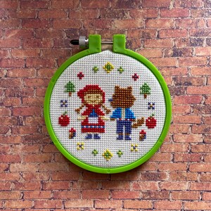 May include: A green embroidery hoop with a white fabric inside. The fabric has a cross-stitch design of Little Red Riding Hood and a wolf.
