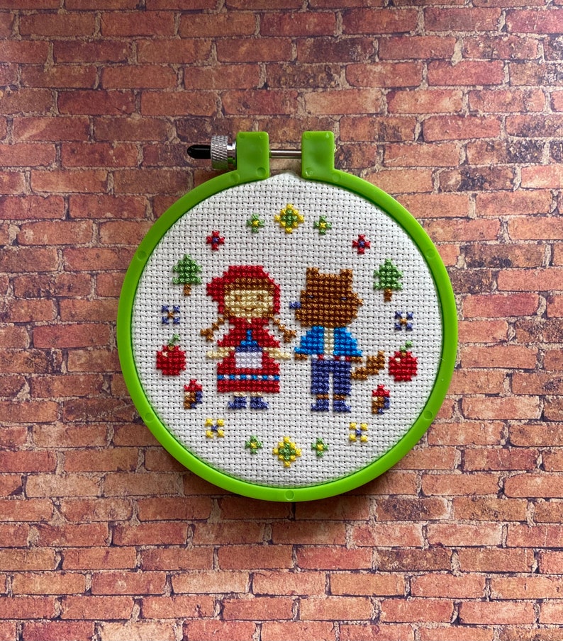 May include: A green embroidery hoop with a white fabric inside. The fabric has a cross-stitch design of Little Red Riding Hood and a wolf.