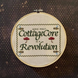 May include: A white embroidery hoop with a cross-stitch design that reads "Done Right Cottagecore is a Revolution". The design features green lettering, red mushrooms, and yellow bees.