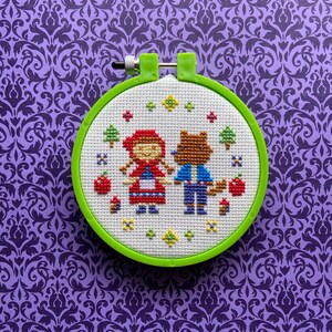 May include: A green embroidery hoop with a white fabric inside. The fabric has a cross-stitch design of Little Red Riding Hood and a wolf.