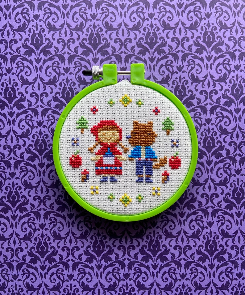 May include: A green embroidery hoop with a white fabric inside. The fabric has a cross-stitch design of Little Red Riding Hood and a wolf.