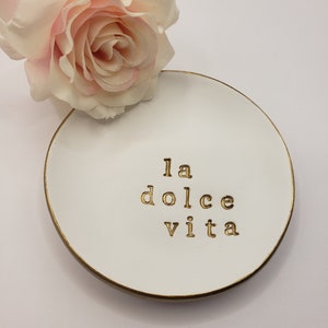 May include: White ceramic trinket dish with a gold rim and the words "la dolce vita" written in gold lettering. A pink rose is sitting on a white surface behind the dish.