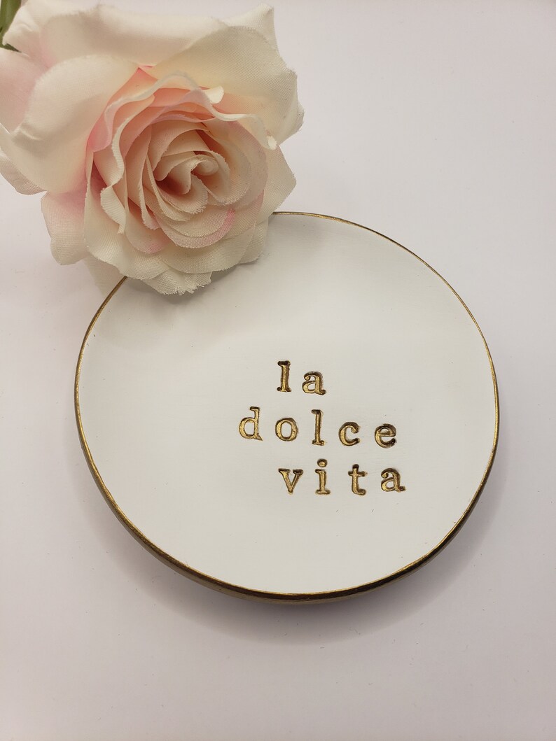 May include: White ceramic trinket dish with a gold rim and the words "la dolce vita" written in gold lettering. A pink rose is sitting on a white surface behind the dish.