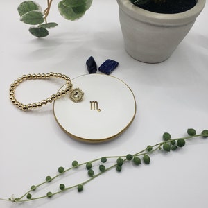 May include: A white ceramic dish with a gold rim and the zodiac symbol for Scorpio. A gold beaded bracelet with a gold charm is on the dish. The dish is on a white surface with green plants.