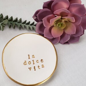May include: White ceramic ring dish with a gold rim. The dish has the phrase "la dolce vita" written in gold lettering.